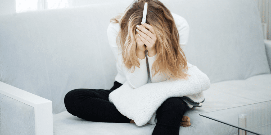 Young woman in black pants and white shirt sets on a white couch holding a white blanket. She is holding a pregnancy test with both hands and has her arms covering her face, appearing to be frustrated