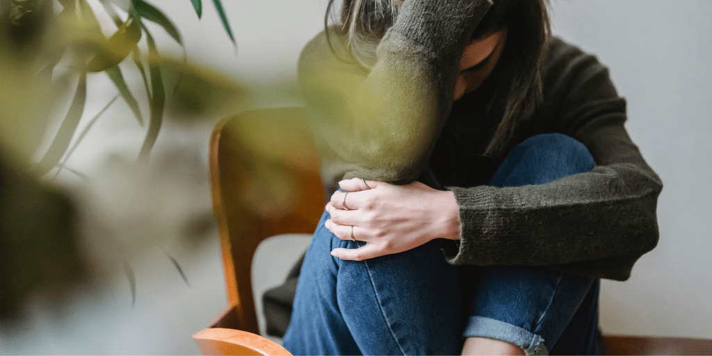 Young woman in jeans and a brown sweater sets with her knees up in a wooden chair, covering her head with her hands, feeling worried