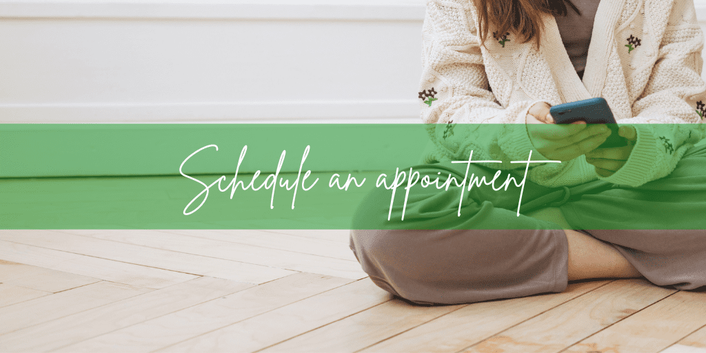 Young woman sets on a wooden floor in lounge-wear and a cardigan searching on her smart phone "Schedule an appointment"