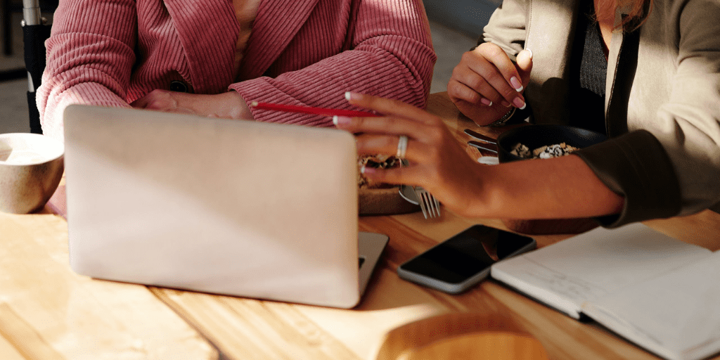 Two women look together at a laptop setting on a table next to notebooks, phones, and lunch