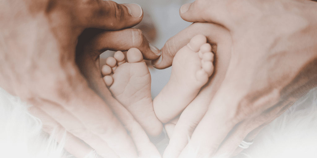 adult male and female hands hold infant feet, making the shape of a heart