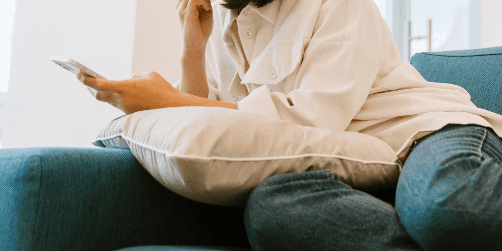 Woman in jeans and white shirt sets on teal couch holding a pillow and her phone