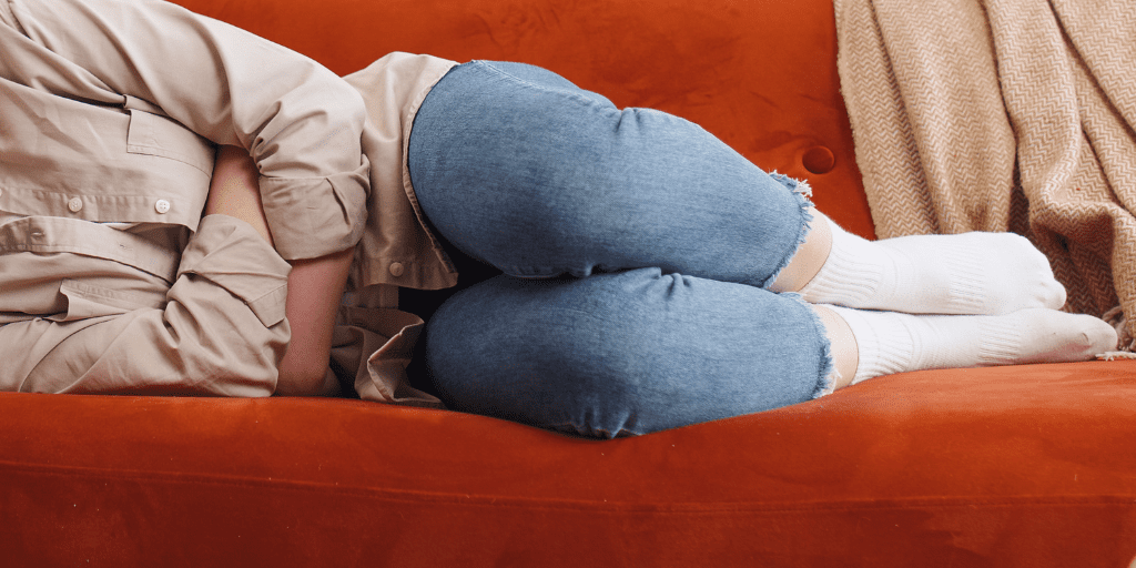 Woman in jeans and tan shirt lays on a red couch holding her abdomen in discomfort