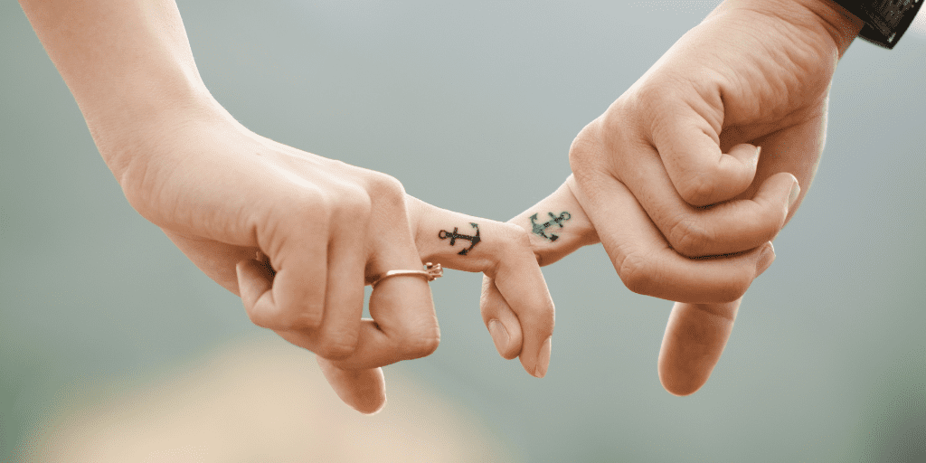 A man and woman are holding hands with just their index fingers interlocked. They have matching tattoos that look like boat anchors.